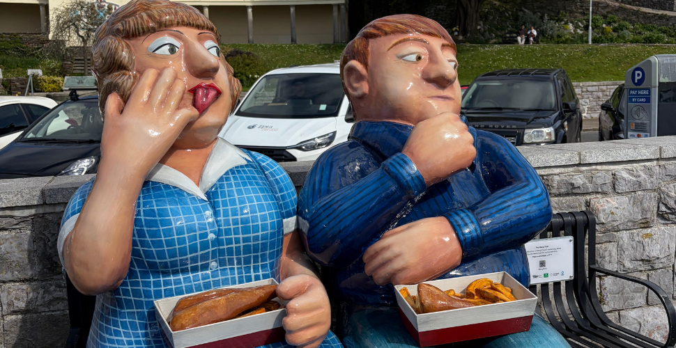 Colourful life-size statues of a man and woman sitting on a bench, each holding a cardboard tray of fish and chips, with parked cars and a grassy area with buildings visible in the background.